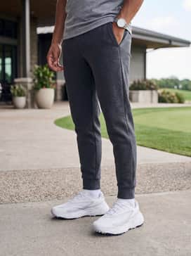 Ua Halo Hybrid Spikeless – A clean athleisure ensemble with charcoal grey tech-fleece joggers, a light grey fitted quarter-zip pullover, and a white performance baseball cap.