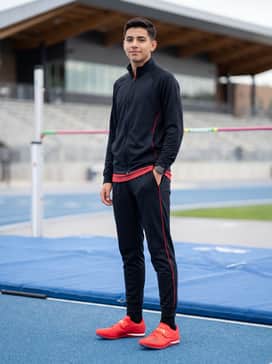 A sleek full-zip track warm-up suit in black with red piping, worn over a moisture-wicking base layer for pre-event preparation.