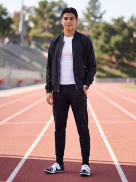 Pre-meet warm-up attire consisting of black tapered tech-fleece joggers, a white moisture-wicking compression shirt, and a gold-accented windbreaker for a unified look.