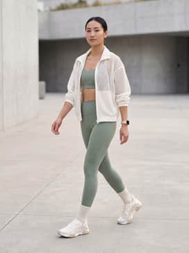 Ua Sculpt Trainer – High-waisted compression leggings in sage green paired with a matching sports bra and a white oversized mesh zip-up jacket for a sleek gym look.