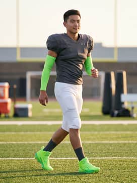 A full competitive uniform featuring a charcoal grey team jersey, white integrated padded football pants, and neon green compression arm sleeves to match the cleats.