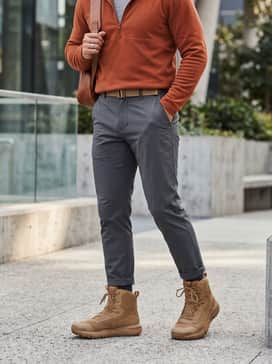 A modern Gorpcore ensemble consisting of graphite grey technical hiking trousers, a burnt orange quarter-zip fleece, and a charcoal high-performance rain shell.