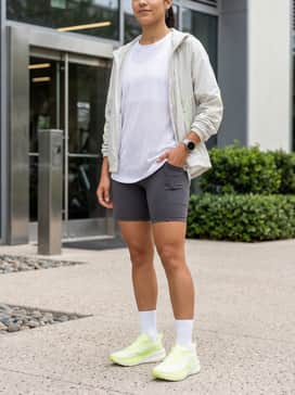 Ua Velociti Spd 5 – Charcoal grey biker shorts combined with an oversized white mesh t-shirt and a lightweight neon-accented windbreaker for a modern gym ensemble.