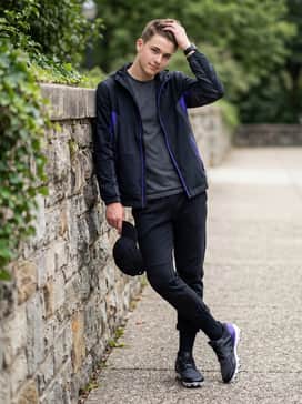 A post-game travel outfit featuring tapered black tech-fleece joggers, a purple-accented windbreaker, and a matching team cap.