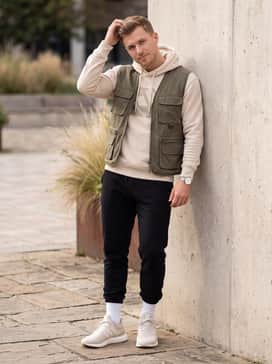 Black tapered joggers combined with a graphic hoodie and an olive green utility vest, finished with white crew socks pulled over the hems for a modern streetwear aesthetic.