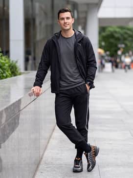 Voltra Mens Sneakers – A high-performance athleisure outfit pairing black moisture-wicking track pants with silver side-striping, a fitted black compression top, and a lightweight weather-resistant windbreaker.