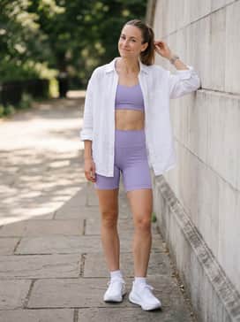 A summer fitness outfit pairing lavender high-waisted biker shorts with a matching sports bra and an oversized white linen shirt worn open for breathability.