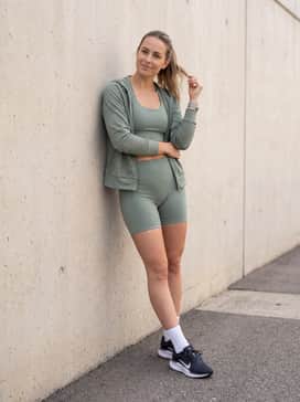 Winflo 11 – Sage green seamless biker shorts and a matching longline sports bra topped with a mesh zip-up hoodie for a modern gym look.