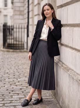 Womens Grand Ambition Alette Buckle Loafers – A charcoal grey pleated midi skirt combined with a tucked-in cream blouse and a structured black blazer for a polished office ensemble.