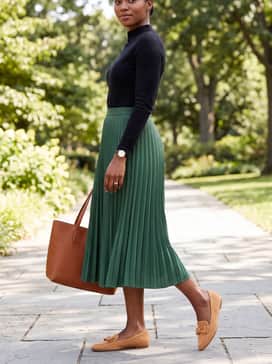 A pleated emerald green midi skirt styled with a slim-fit black mock-neck top and a structured cognac leather tote bag.