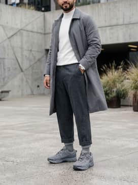 Zoom Vomero Roam – Cropped charcoal wool trousers, a white mock-neck sweater, and a long grey overcoat for a sophisticated, athletic-inspired minimalist look.